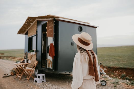 Woman standing by a tiny house on wheels in a rural landscape, capturing the essence of mobile living.