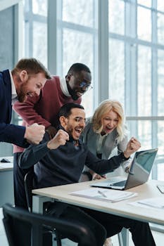 A diverse team in an office celebrates a successful achievement while looking at a laptop.